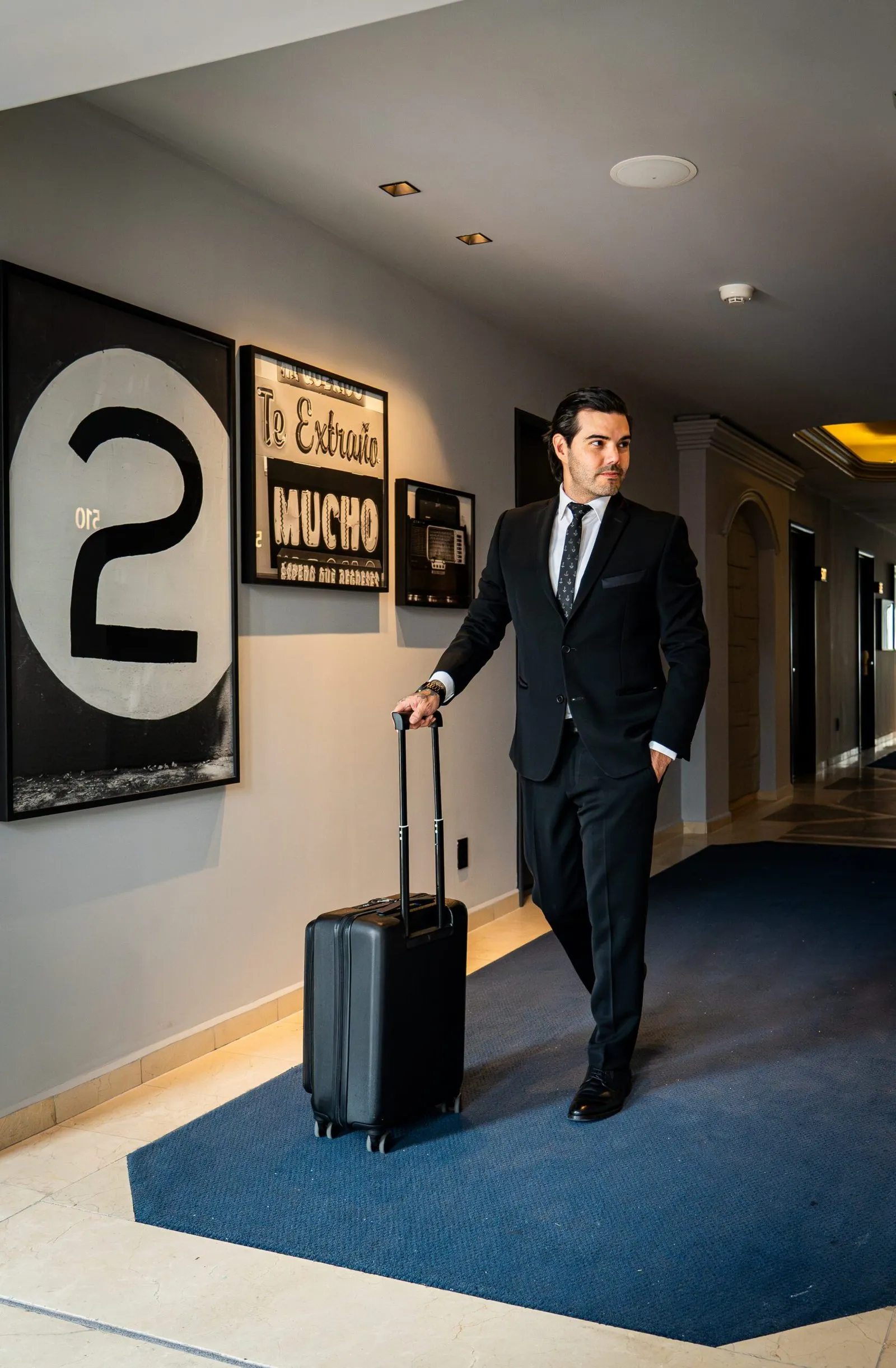 Modelo masculino elegantemente vestido, saliendo del hotel con una maleta, listo para continuar su viaje.